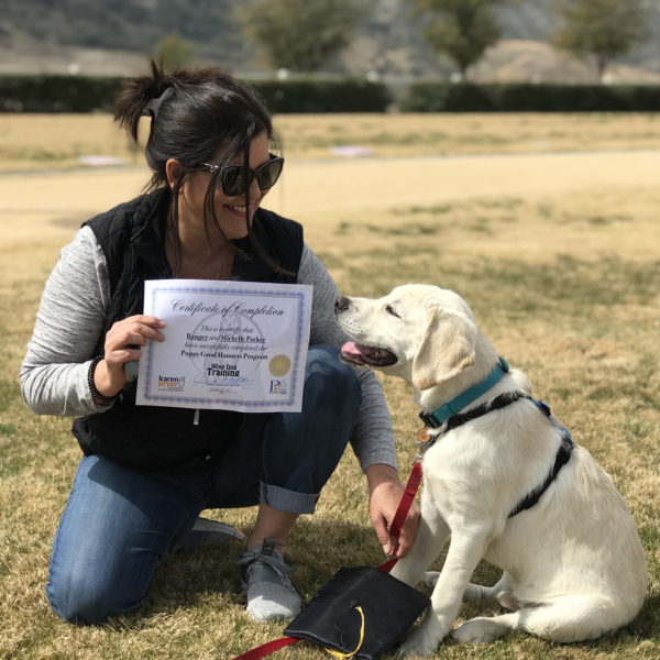 A lady in sunglasses kneeling next to a yellow lab, smiling at each other, showing off their completion certificate